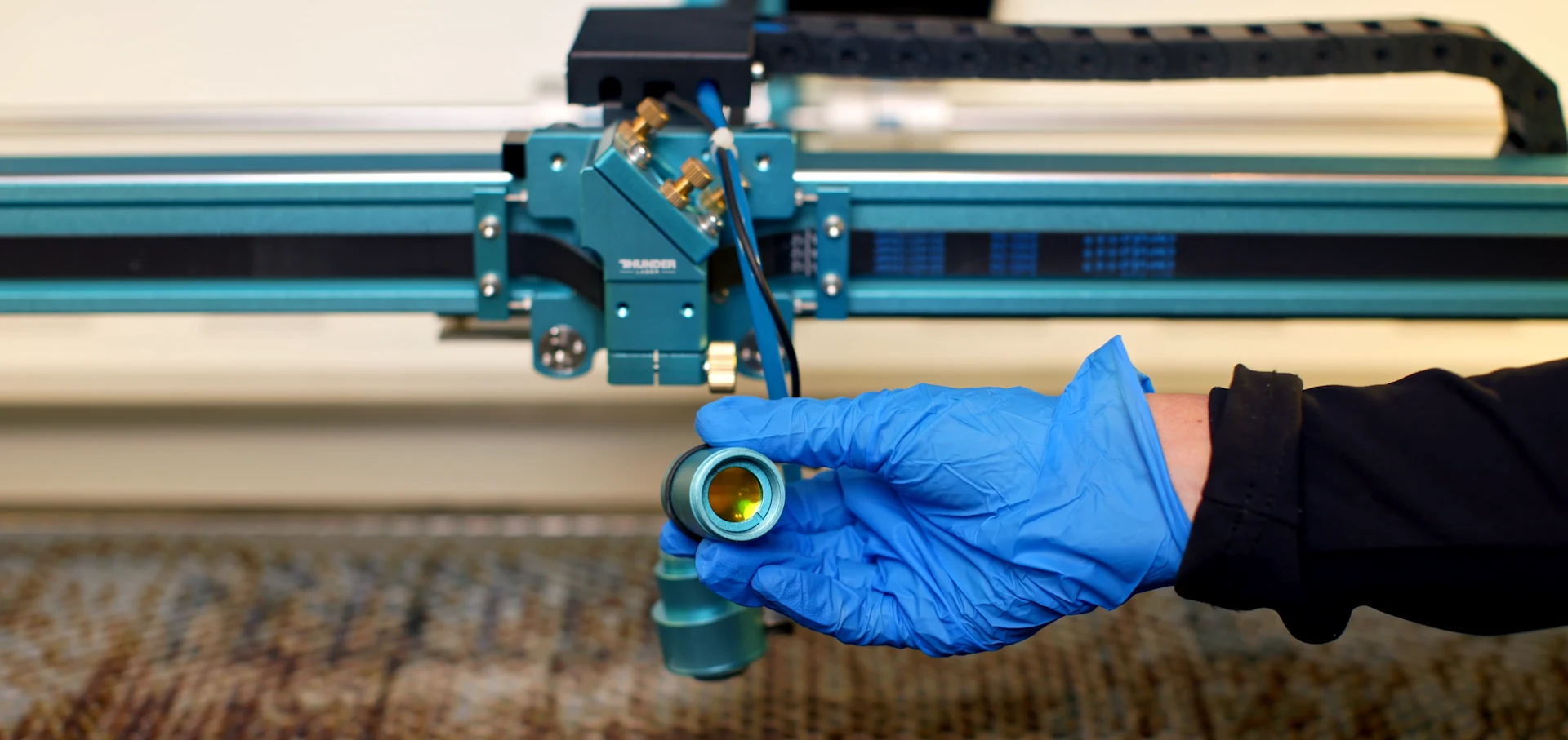 Close-up of a gloved hand holding a laser lens in front of a Thunder Laser machine during lens and mirror maintenance.