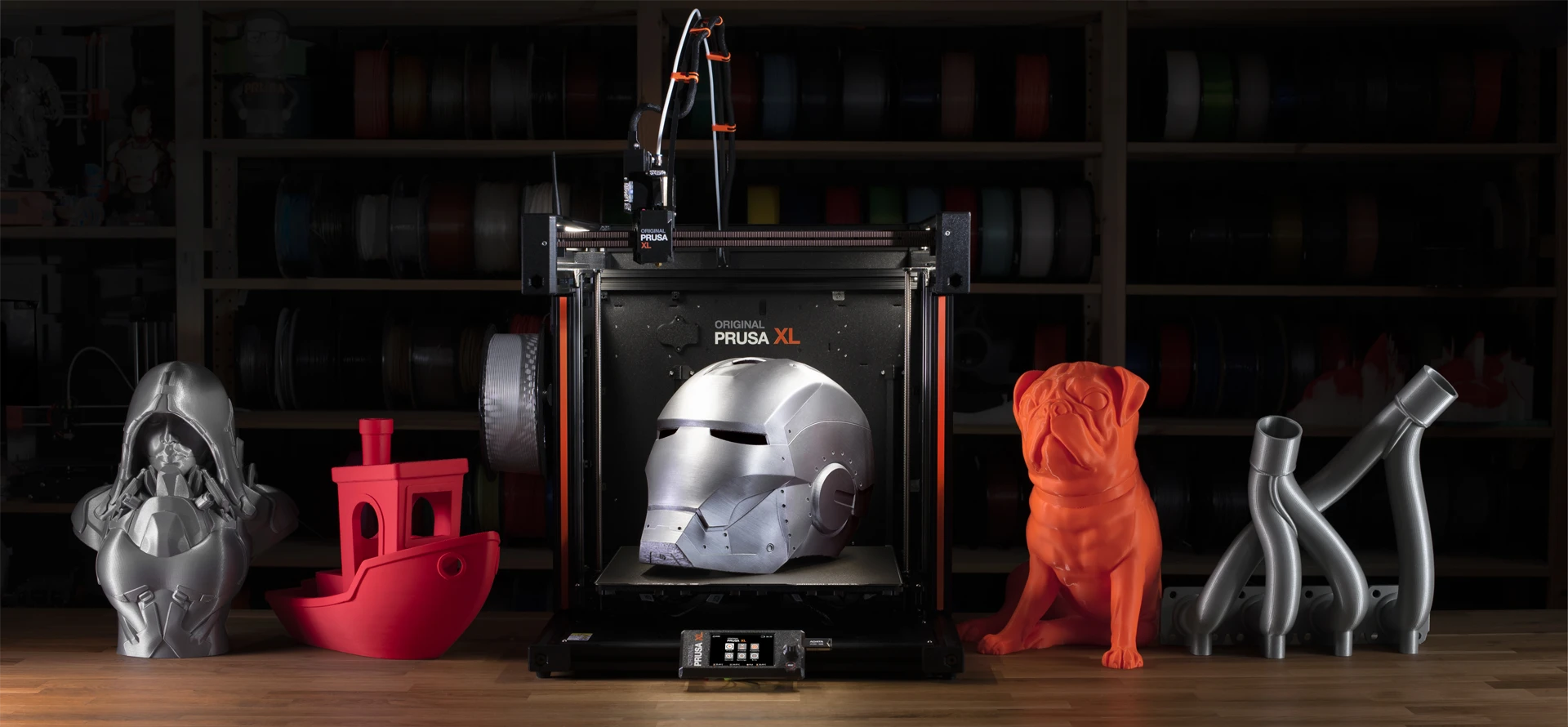 Original Prusa XL 3D printer on a workshop bench surrounded by finished 3D prints, including a metallic helmet, red boat, silver bust, orange dog and curved grey models.
