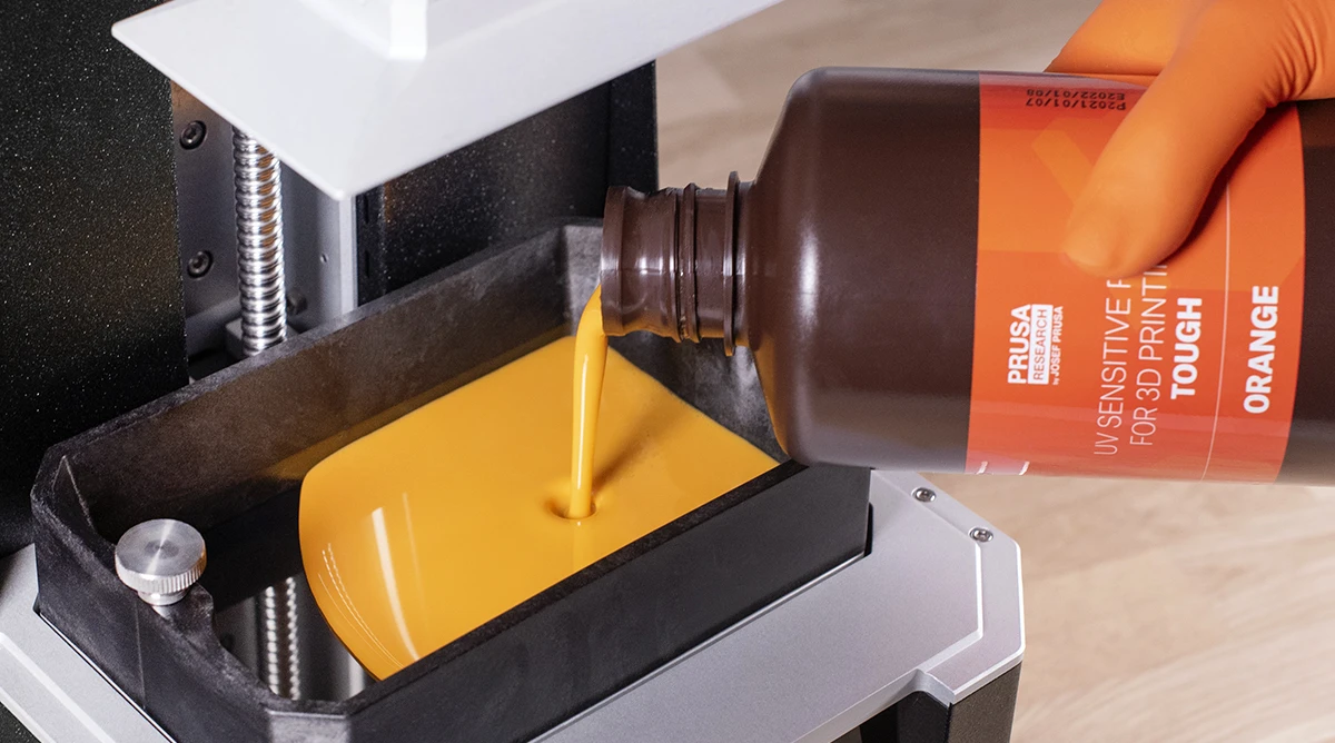 Orange liquid photopolymer resin being poured into the resin tank of a Prusa 3D printer.